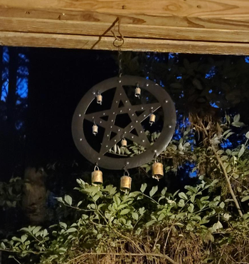 A pentagram wind chime with brass bells hanging from a wooden beam, casting an eerie glow at night.