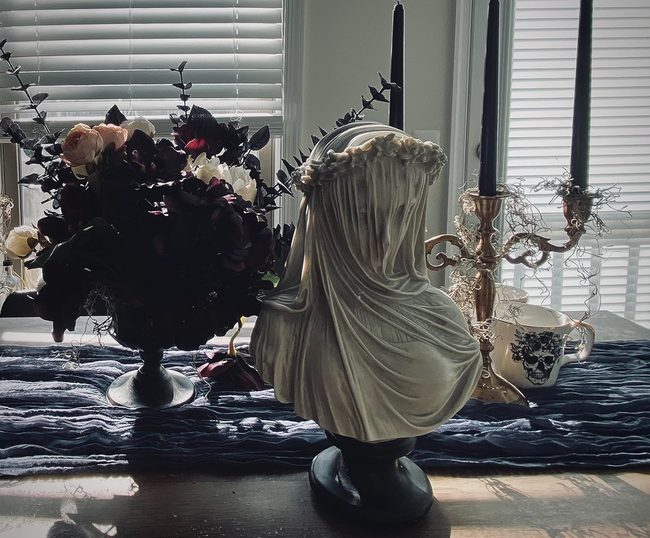 A veiled bust sitting on a table adorned with decorative flowers and a candle holder, radiating a vintage, classic decor vibe.