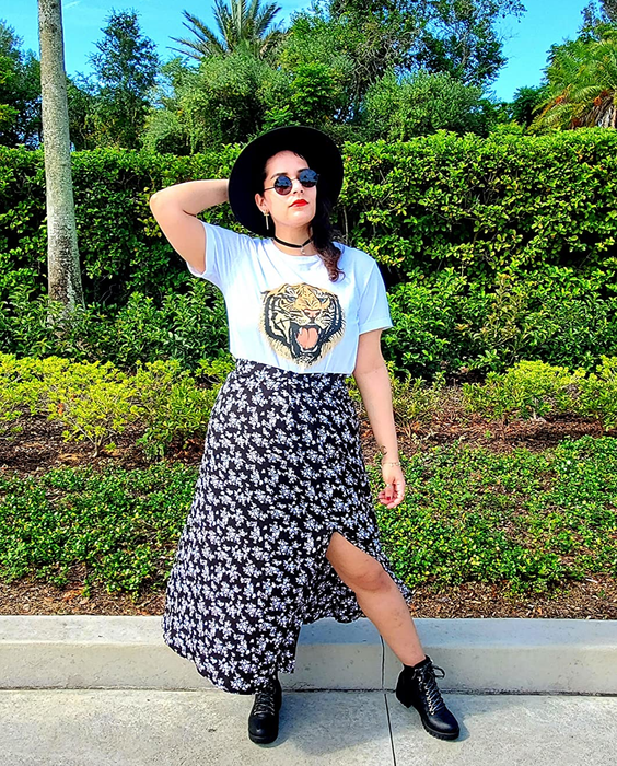 A reviewer styled in a graphic tee, floral skirt, boots, and a wide-brimmed hat, striking a pose with one hand on their hip