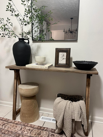 A stylish wooden console table adorned with vases, bowls, and other decor, complemented by a stool and blanket beneath, creating an inviting atmosphere.