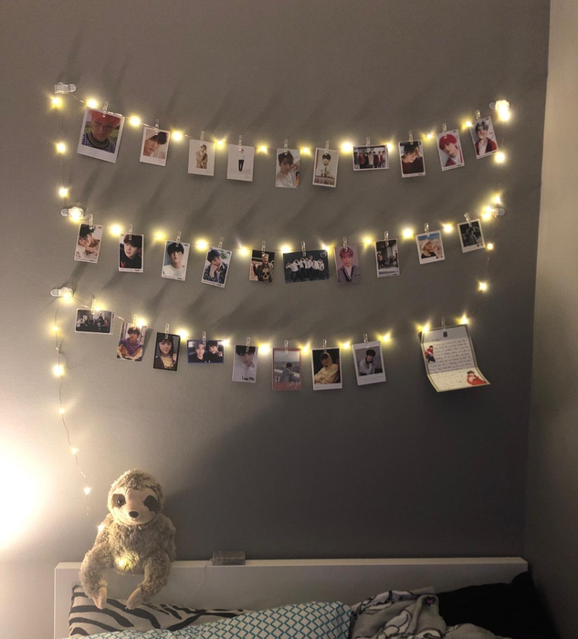 A cozy bedroom with string lights illuminating Polaroid photos on the wall, above a bed adorned with pillows and a plush sloth toy.