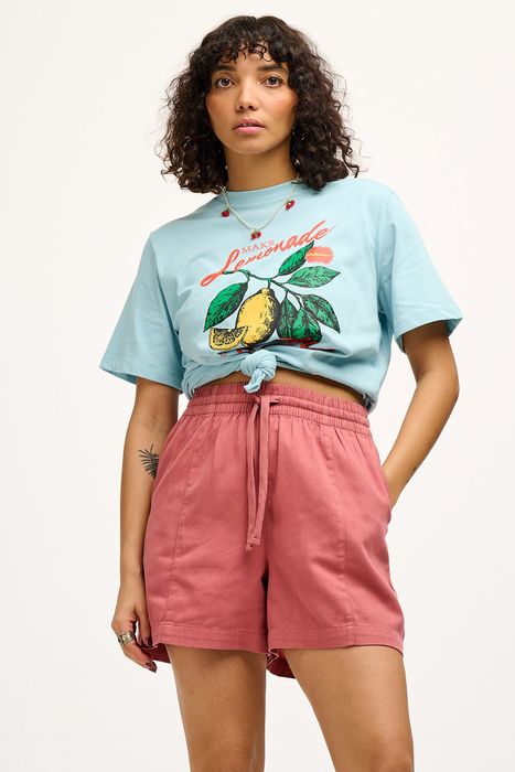 A model casually standing in a tied-up graphic tee and pink shorts.