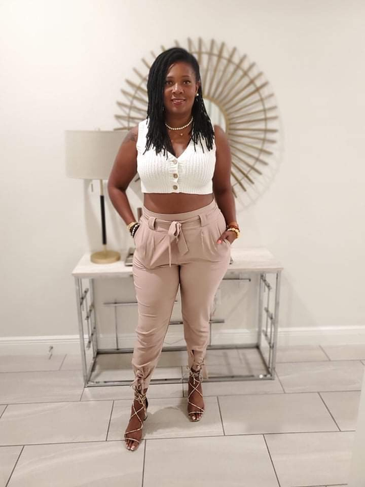 A woman in a white crop top and beige trousers with lace-up heels stands indoors.