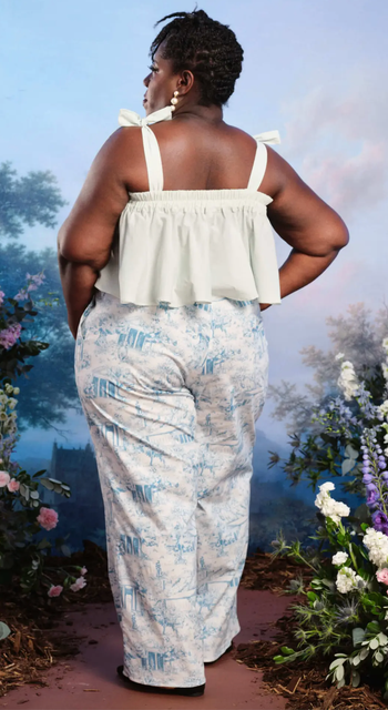 A model stands in a garden wearing high-waisted printed trousers and a ruffled off-the-shoulder top, looking effortlessly chic.