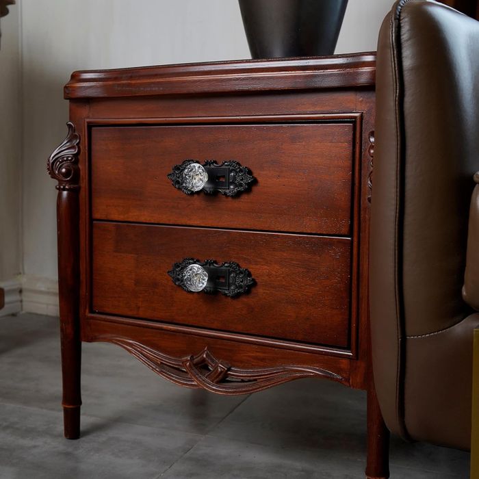 A wooden bedside table featuring two drawers and decorative, ornate handles.