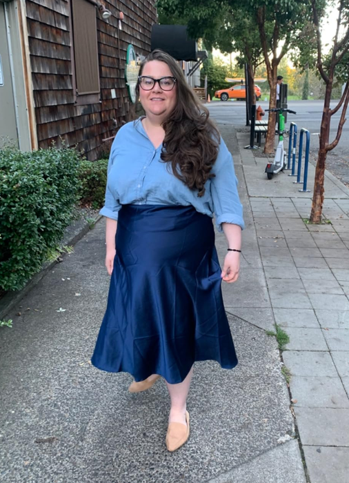 A reviewer wearing a buttoned shirt and pleated skirt, paired with flats, strolling down the sidewalk.