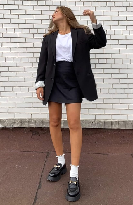 A reviewer in an oversized blazer and shorts, casually leaning against a wall, looking up, with chunky shoes on.