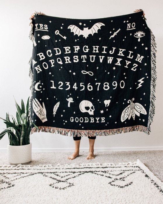 A black blanket featuring an illustrated Ouija board with tassels along the edges.