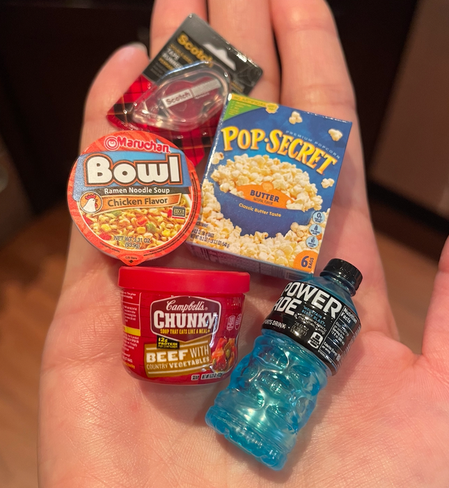 A hand holding five miniature items: Maruchan ramen cup, Pop-Secret popcorn box, Campbell's Chunky Beef with Vegetables soup, and a blue Powerade bottle.