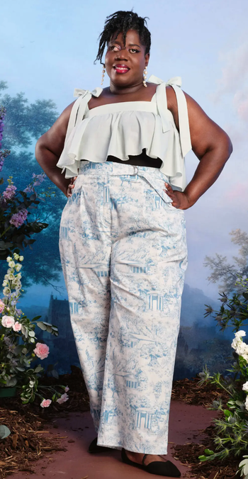A model poses in a ruffled top paired with printed trousers, with a floral backdrop setting the scene.