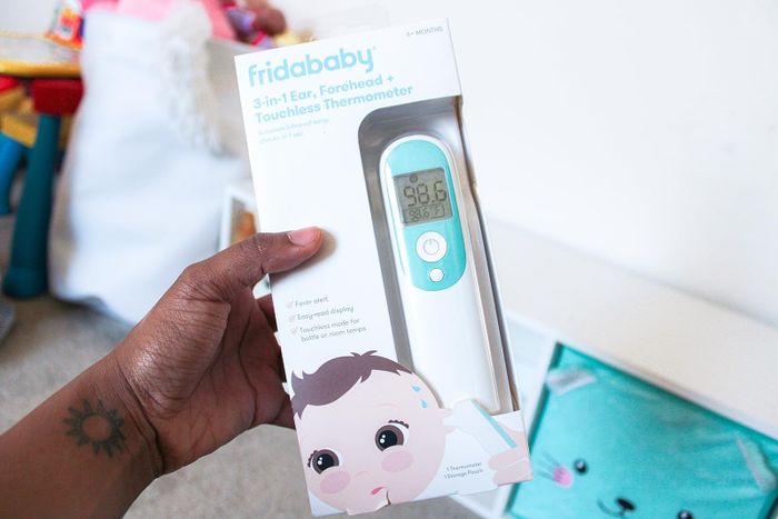 A hand holding the packaging of the Fridababy 3-in-1 thermometer, displaying the device designed for ear, forehead, and touchless temperature measurements.