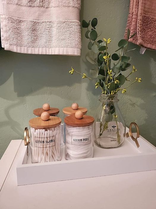 A reviewer’s photo of labeled glass jars with wooden lids, filled with q-tips and cotton pads, arranged on a tray alongside a vase of eucalyptus.