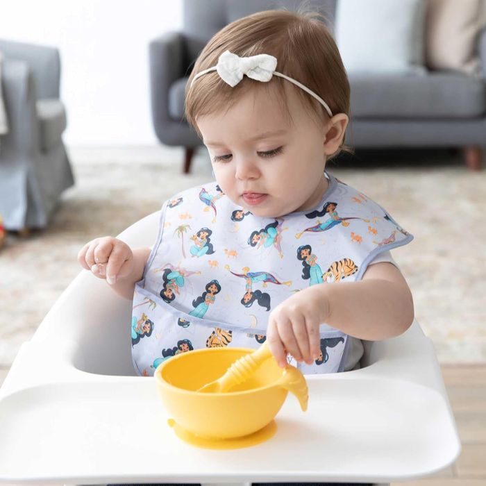 A young child, dressed in a Disney princess-themed bib, plays with a yellow bowl and spoon while seated in a high chair, showcasing baby care and feeding products.