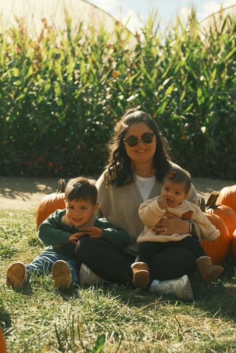 A woman enjoys a pumpkin patch outing with her two young children, all smiling in front of a cornfield—capturing a joyful family moment.