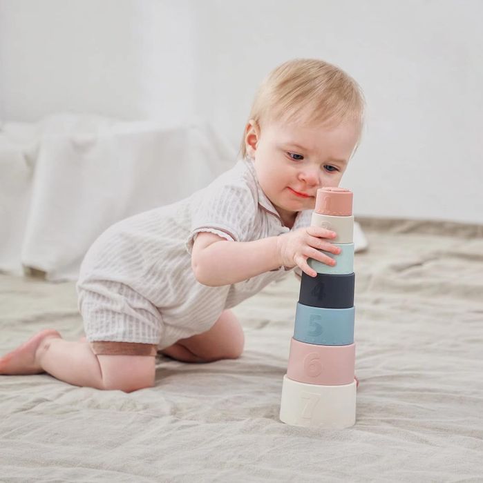 A baby playing with stackable toy cups, numbered from 1 to 7, on a soft surface.