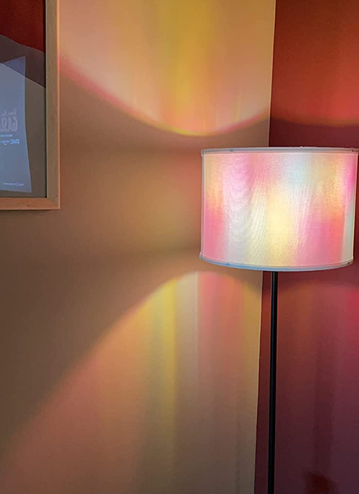 A floor lamp projects a vibrant array of colors onto a wall and surrounding surfaces, with a framed object hanging nearby.