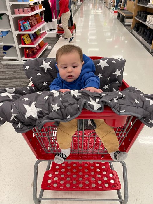 A baby seated in a grey shopping cart cover adorned with white stars.
