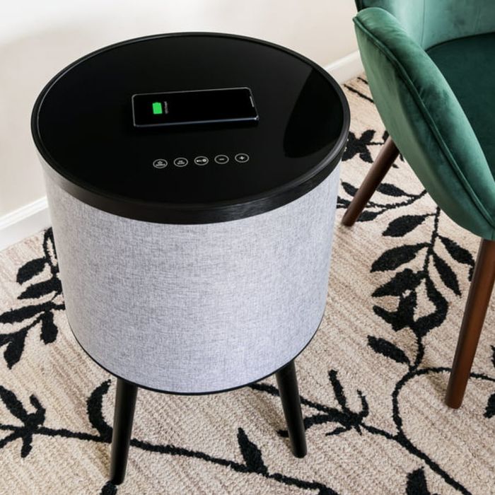 A smart speaker with a digital display and fabric cover, placed on a carpet next to a green chair.