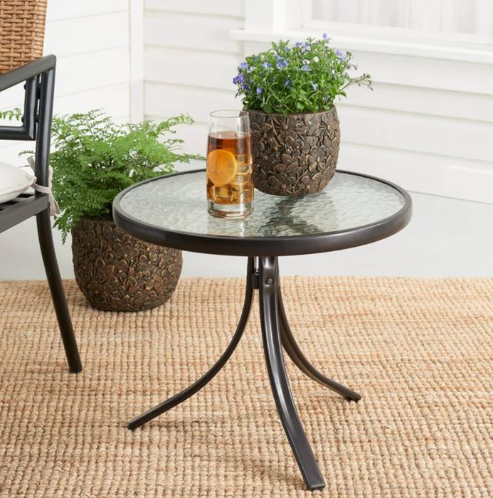 A round glass-top table placed outdoors, with two decorative potted plants on top, set on a textured rug for an added touch of coziness.