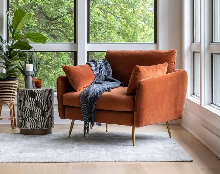 A chic room featuring a trendy burnt orange chair, a geometric side table, and a cozy gray throw, with large windows offering a leafy view
