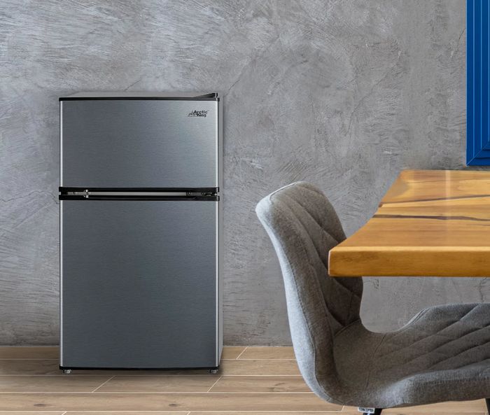 A gray mini fridge placed next to a wooden table and a gray chair in a room with a sleek concrete wall in the background.