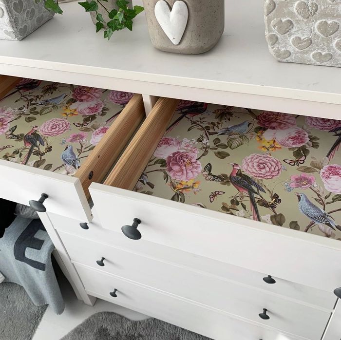 A partially opened drawer showcasing a liner with a floral and bird motif inside a white dresser featuring silver knobs.