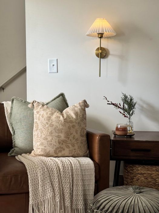 A warm and inviting nook featuring a leather couch, accent pillows, a cozy throw, a wall-mounted lamp, a side table, and a potted plant