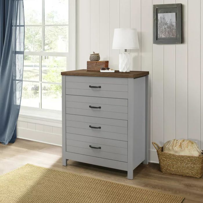 A stylish gray four-drawer dresser with a rich brown top, adorned with a lamp and decorative pieces, placed next to a window in a room.