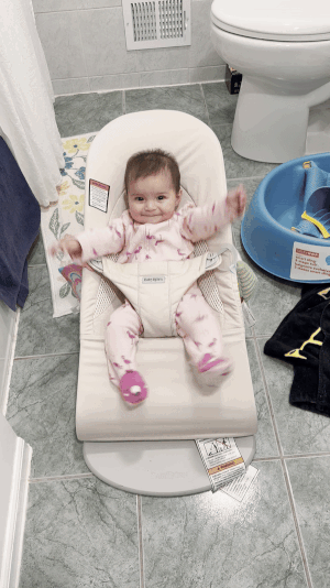 A baby grinning happily while seated in a baby bouncer, set in a bathroom environment with a joyful expression.