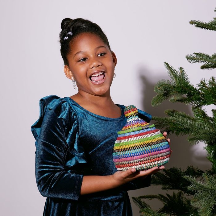 A young girl wearing a dark blue velvet dress holds a brightly colored basket beside a Christmas tree