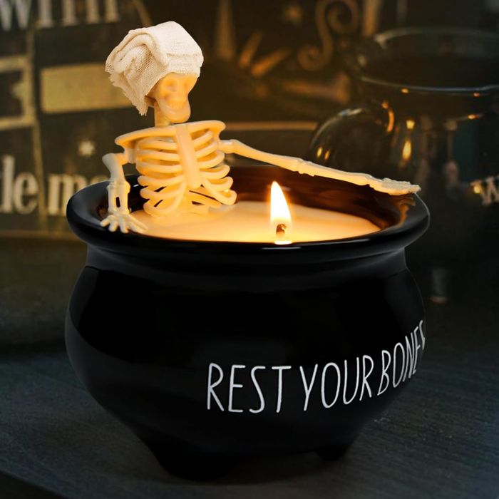 A skeleton figure wrapped in a towel sits in a black cauldron-shaped candle with the words "REST YOUR BONES" on it