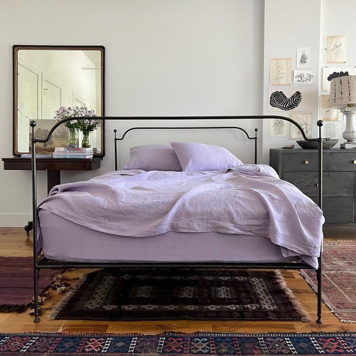 A beautifully made bed featuring an iron frame, fresh purple linens, surrounded by artwork and a floor mirror.