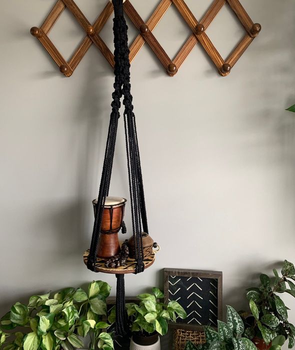 A macrame plant hanger holding a potted plant, set against a wall featuring a diamond-patterned rack with plants on the side.