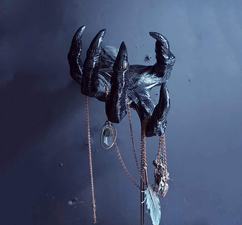 a black wrinkled hand with long, thick nails mounted on a wall, holding pieces of jewelry