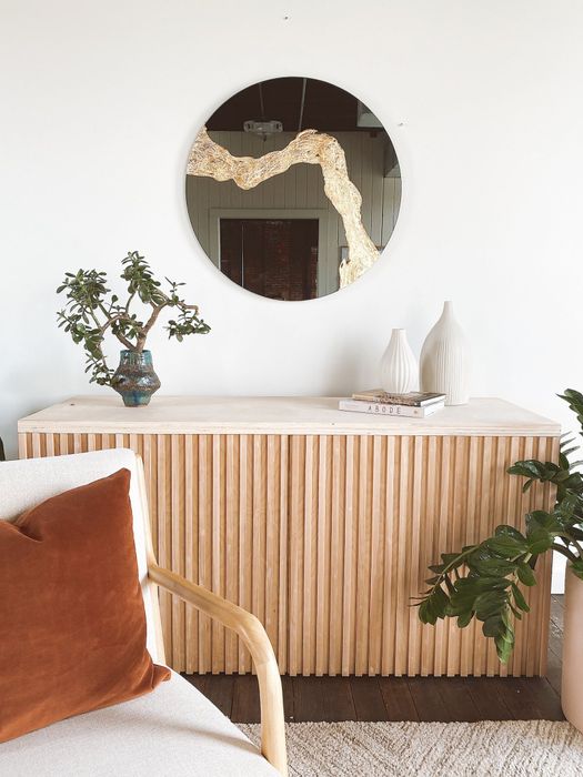 A minimalist room showcasing a wooden sideboard adorned with decor, a round mirror with a gold accent above it, and a chair with an orange cushion