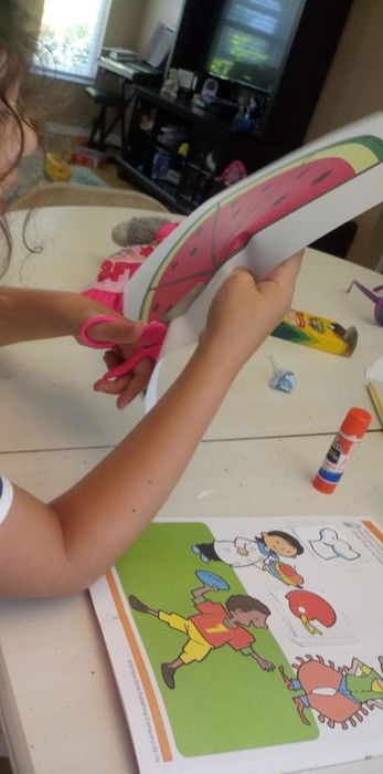 A reviewer's child carefully cutting out a watermelon illustration from the activity pad using the yellow scissors