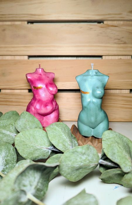 Two candles sculpted like torsos, resting on a wooden surface surrounded by fresh green leaves.