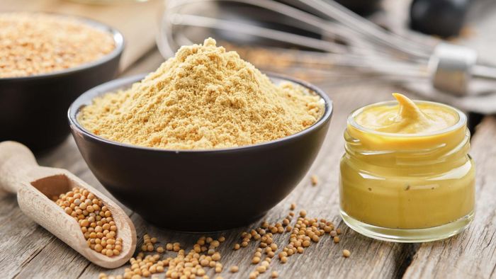 A bowl of mustard powder placed next to a jar of mustard.