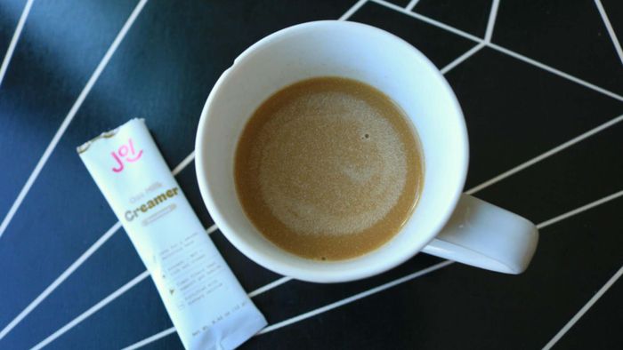 A cup of coffee with an unopened Joi Oat Milk sachet beside it.