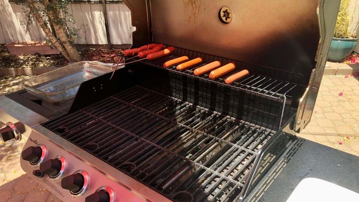 Several hot dogs rest on the top rack of an open grill under the bright sun.