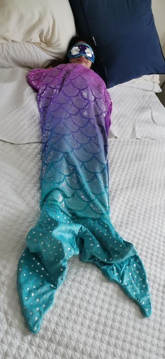 A child is shown wearing a mermaid tail blanket and a sleep mask, enjoying a cozy moment.
