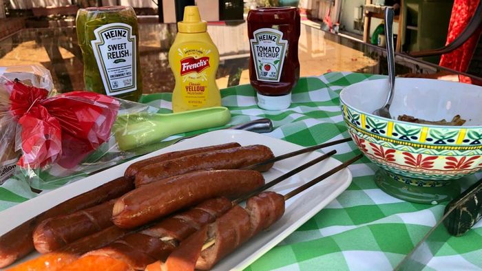 Grilled hot dogs are arranged on a plate, with a spread of condiments and toppings laid out on the table beside them.