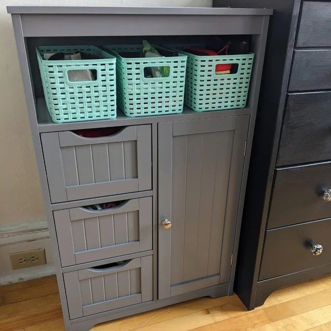 A freestanding gray cabinet featuring three drawers, one door, and a rack that holds three baskets for added storage.