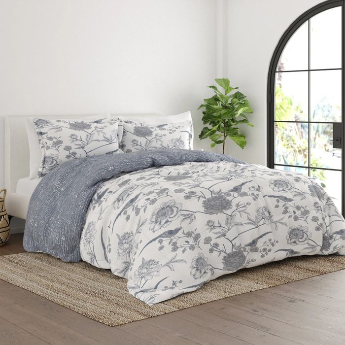A white and gray comforter adorned with floral and bird patterns, complemented by matching pillowcases, resting on a brown rug and hardwood floor.