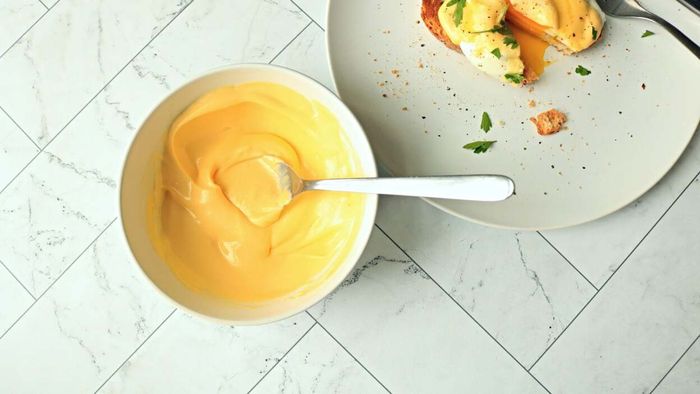 A serving of hollandaise sauce rests beside a plate with an egg on toast.