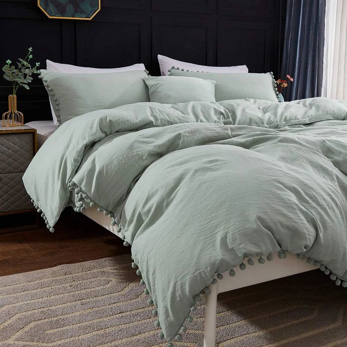 Sage green duvet cover with pom-pom fringe and matching pillowcases resting on a light wooden bed frame against a black wall.