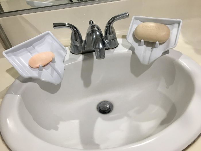 Two white soap dishes positioned on the side of the reviewer's sink. They're angled downward, featuring small poles to secure the soap in place, as well as a channel to direct residue downward.