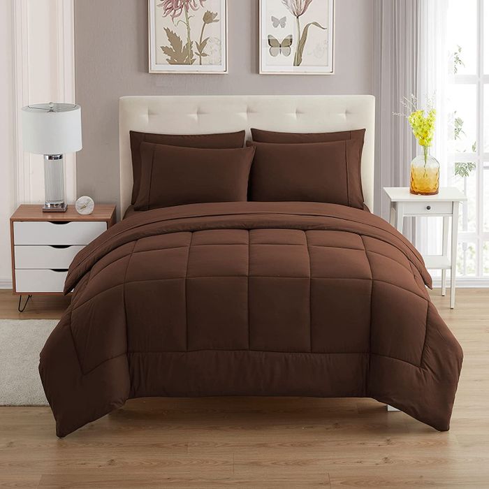 A front view of a brown comforter on a white tufted bed frame, next to a wooden side table with a white lamp.