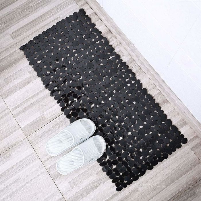 A lifestyle photo of the black pebble bath mat placed in front of a shower, with two white slippers on top of it.