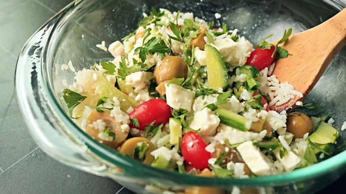 A refreshing rice salad featuring cucumbers, tomatoes, olives, parsley, and feta cheese, all tossed together in a bowl.